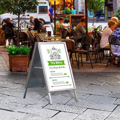 A silver framed A-Board advertising the Tea Menu outside of a coffee shop with tables all around