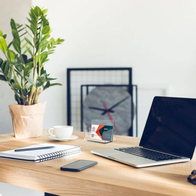 Business card holder on top of a desk that also has a laptop, a notebook, a plant, a phone, and a mug on it