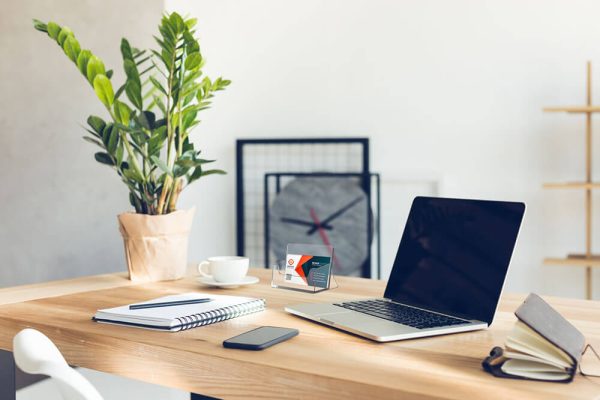 Business card holder on top of a desk that also has a laptop, a notebook, a plant, a phone, and a mug on it