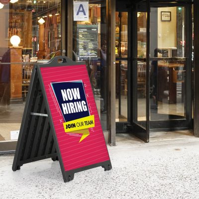 Now hiring sign on a Black A-Board SignPro Sidewalk sign outside of a small cafe with a revolving door