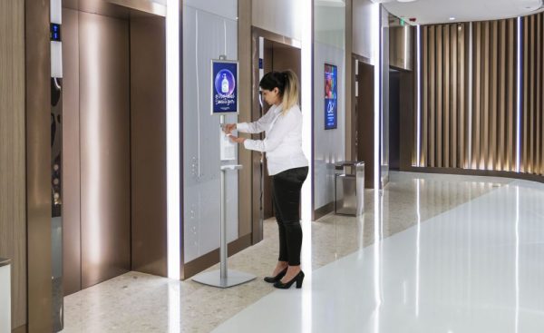 woman applying hand sanitizer from a floor standing dispenser in the hallway of a hospital
