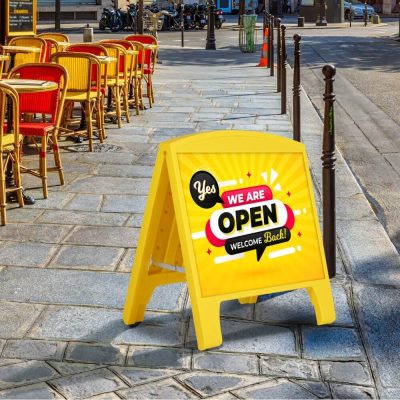 A yellow sandwich board outdoor sidewalk sign holder in front of an outdoor seating area that reads 